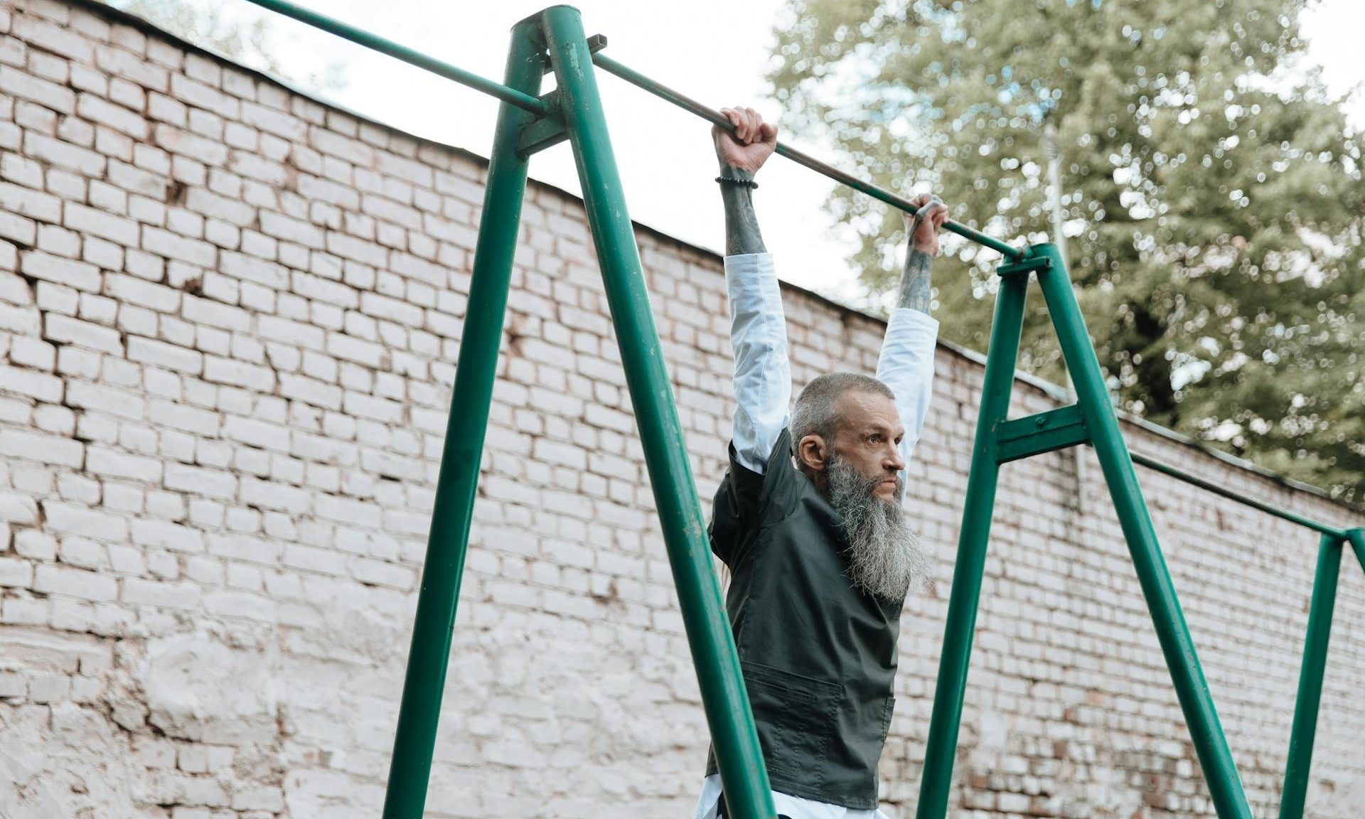 man working out dead hang outside on pull up bar older man