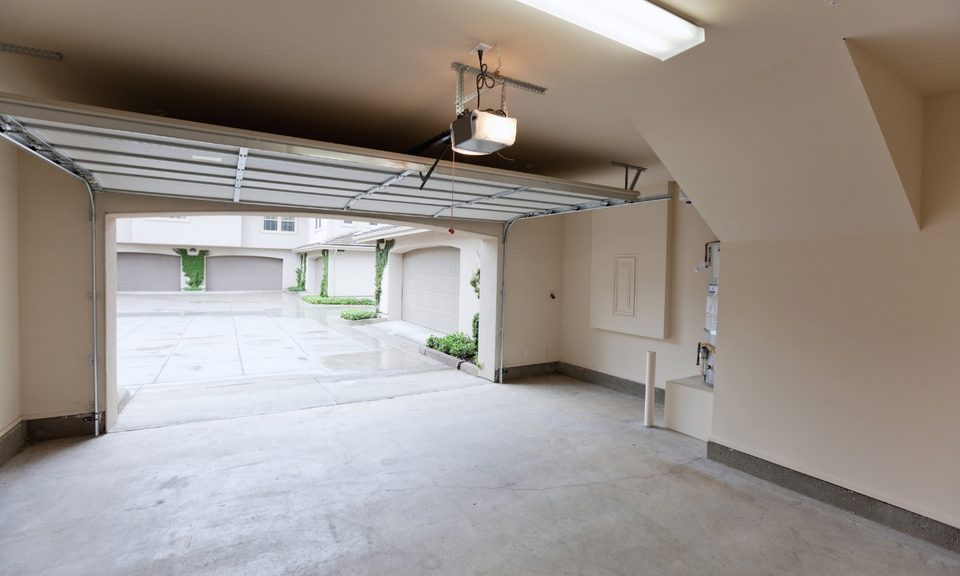 The inside of a garage with beige/cream colored walls with a white garage door folded up to offer a view of other neighboring houses