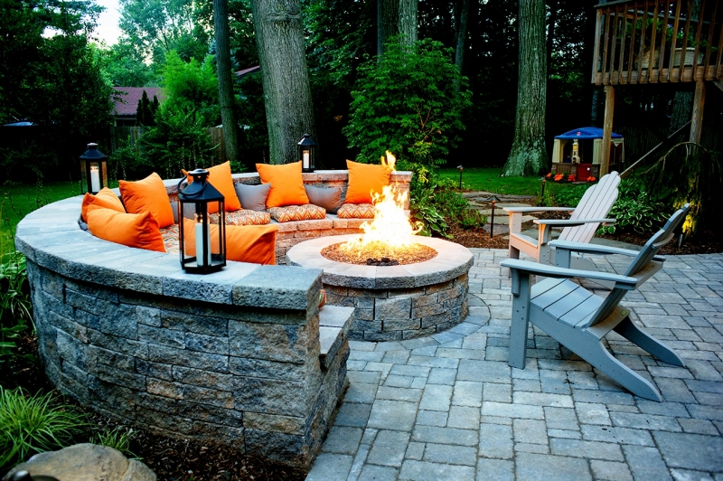 A stone fire pit with orange cushions and lanterns on a patio, with a chair and greenery in the background.