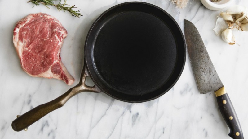 Cast iron skillet on marble slab with steak and knife.