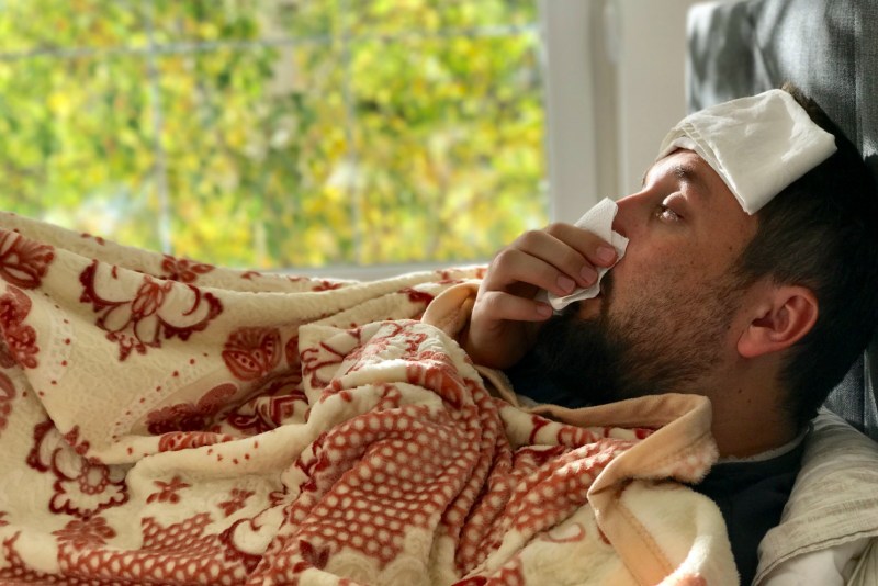 Man sick in bed, blowing his nose into a tissue.