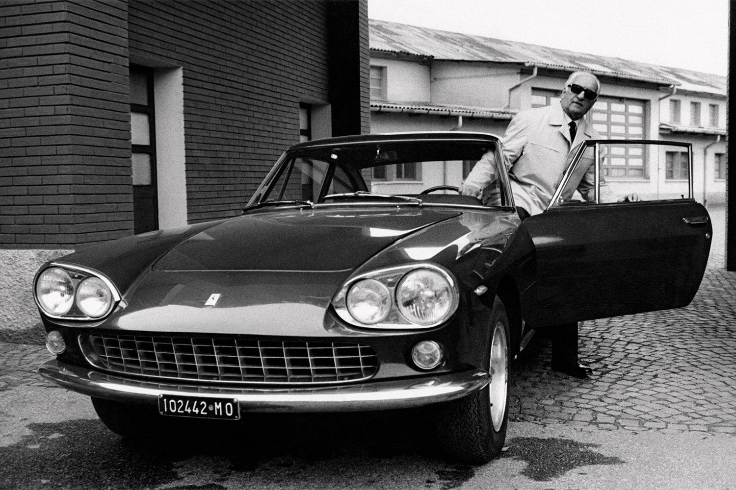 Enzo Ferrari getting out of a Ferrari in front of brick buildings.
