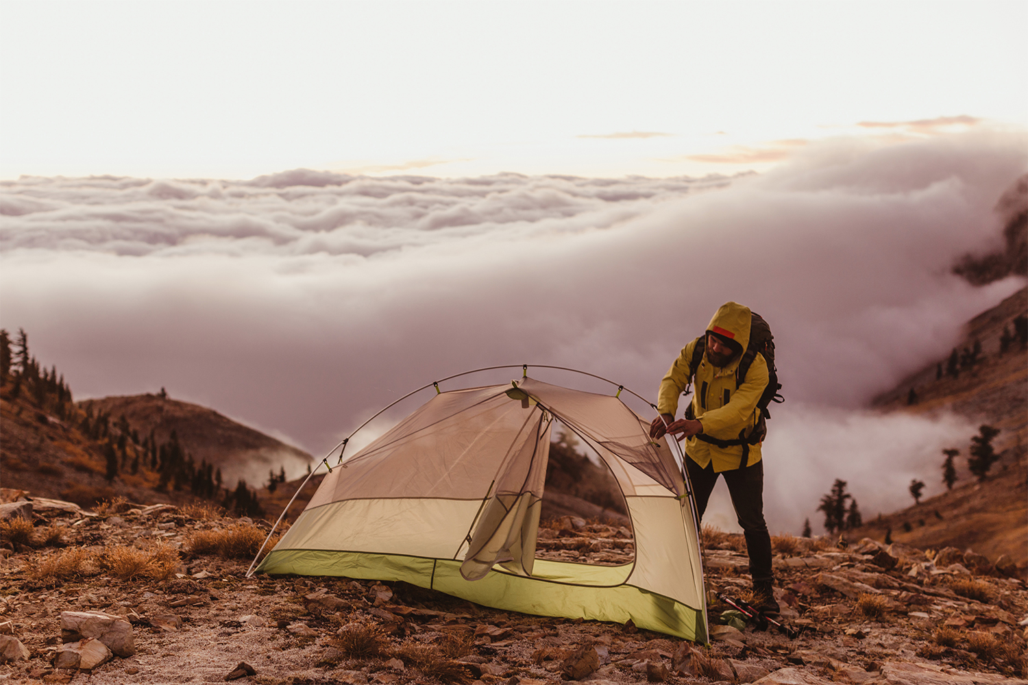 A man wearing a hooded jacket attempting to set up a tent in the wilderness.