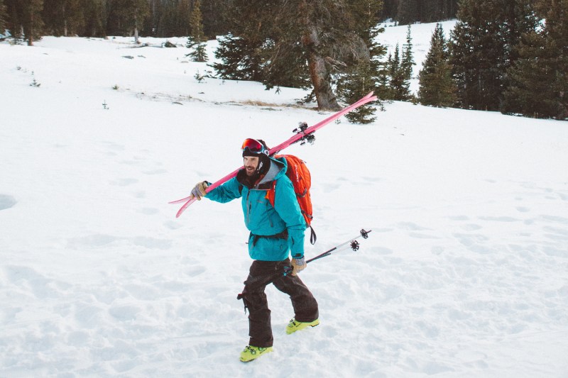 Man trekking through snow carrying skis and ski poles.