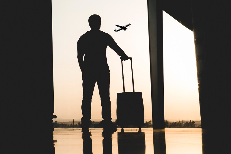 Man with a suitcase watching an airplane take off through the airport window.