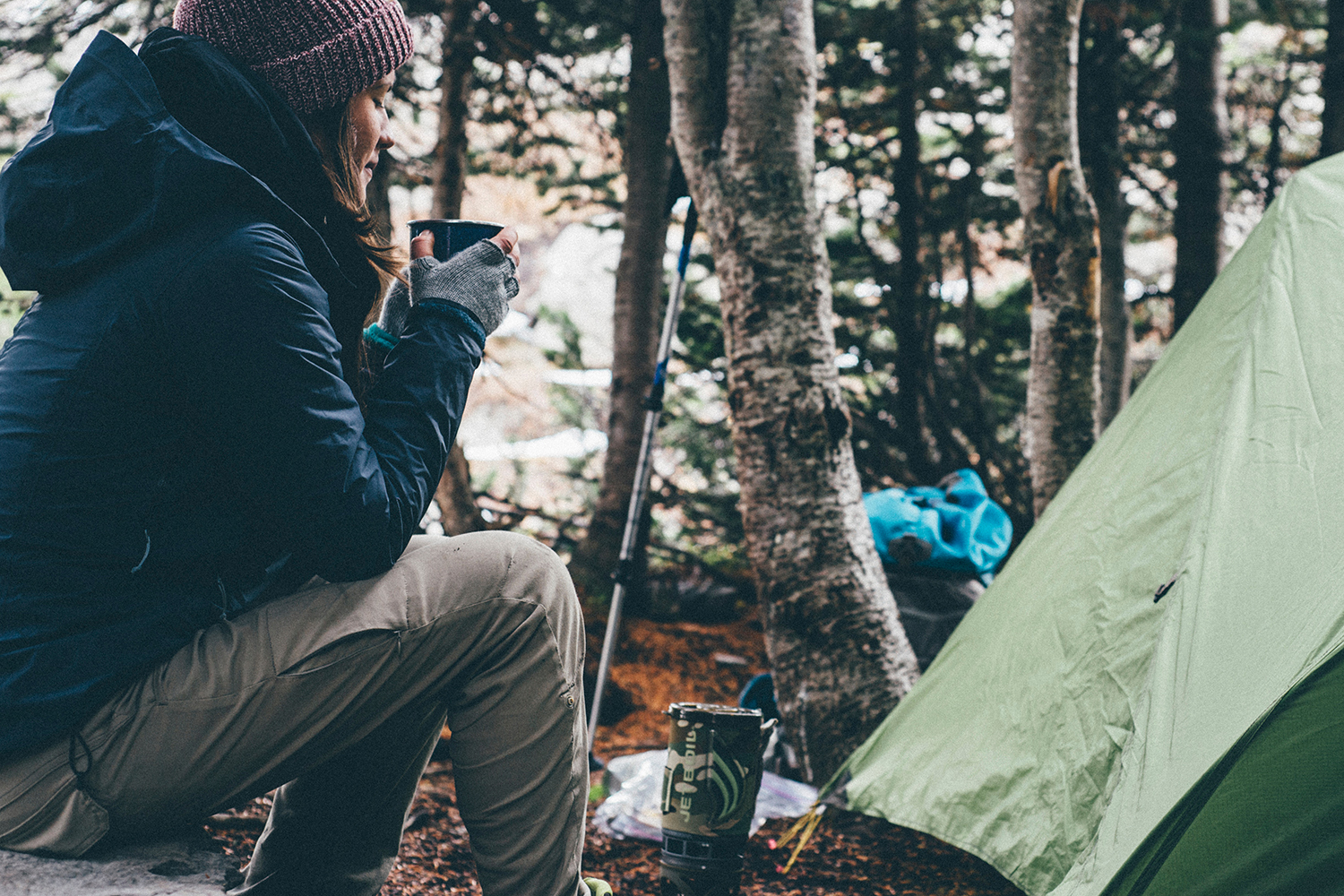 A woman sitting and drinking a warm beverage by a green tent.