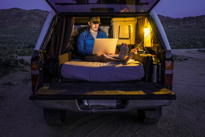 Man relaxing with a laptop in the back of a truck on a mattress.