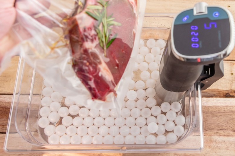 A hand holding a vacuum-sealed bag containing meat on a tray with a sous vide machine.