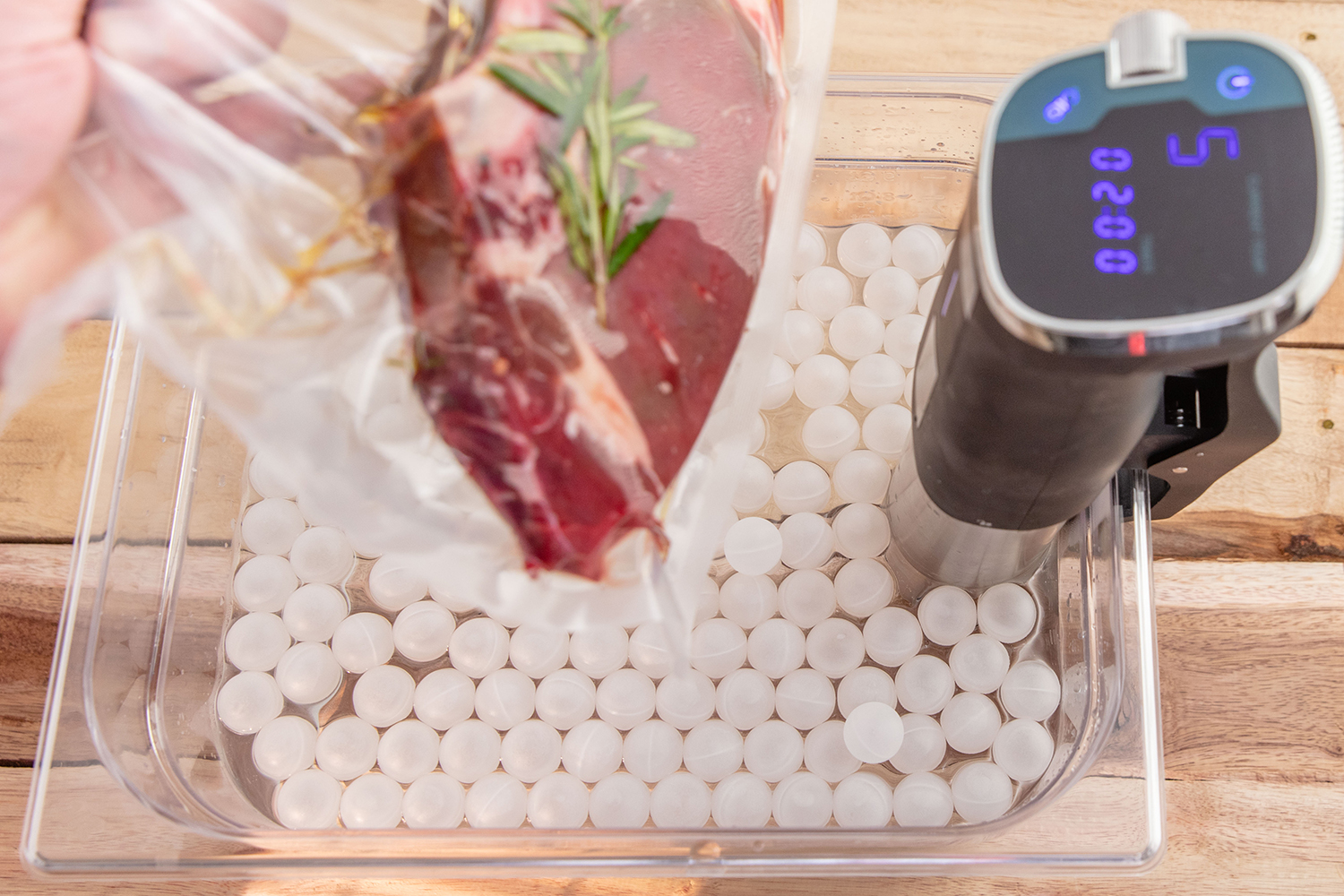 A hand holding a vacuum-sealed bag containing meat on a tray with a sous vide machine.