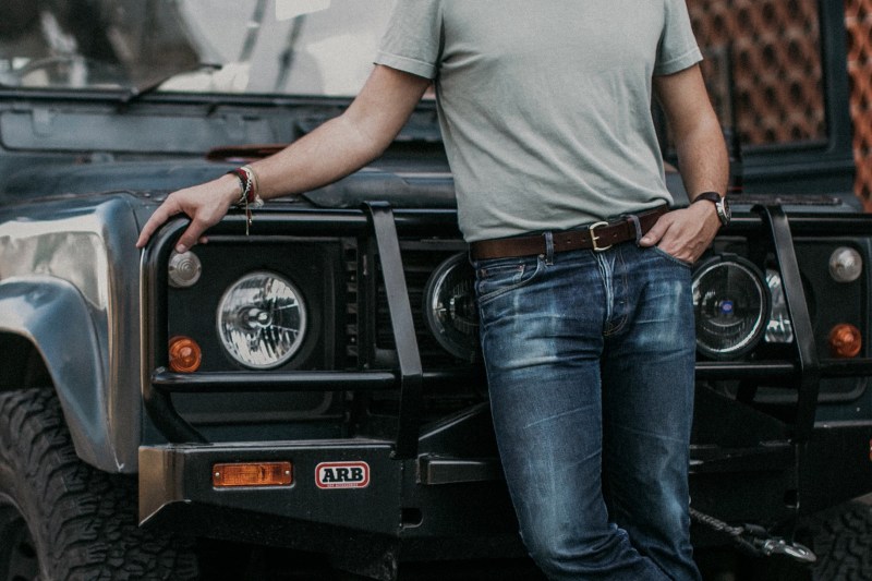 Torso shot of a man wearing jeans and a leather belt while holding onto his vehicle.