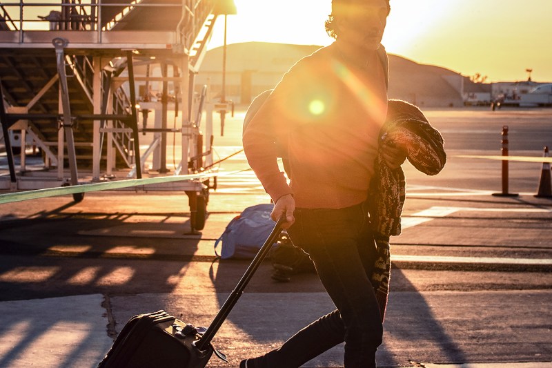 Traveler pulling a rollaboard suitcase outside an airport, sun shining directly into the camera.