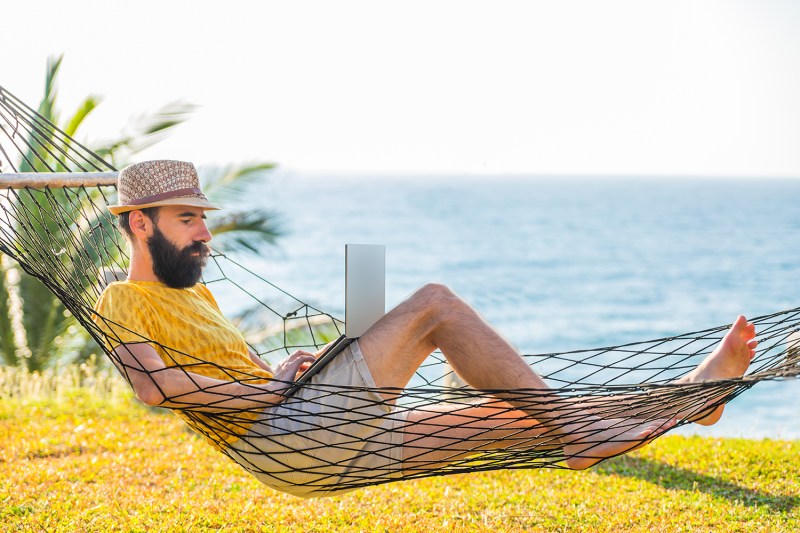 Man working on a laptop from a hammock by the ocean.