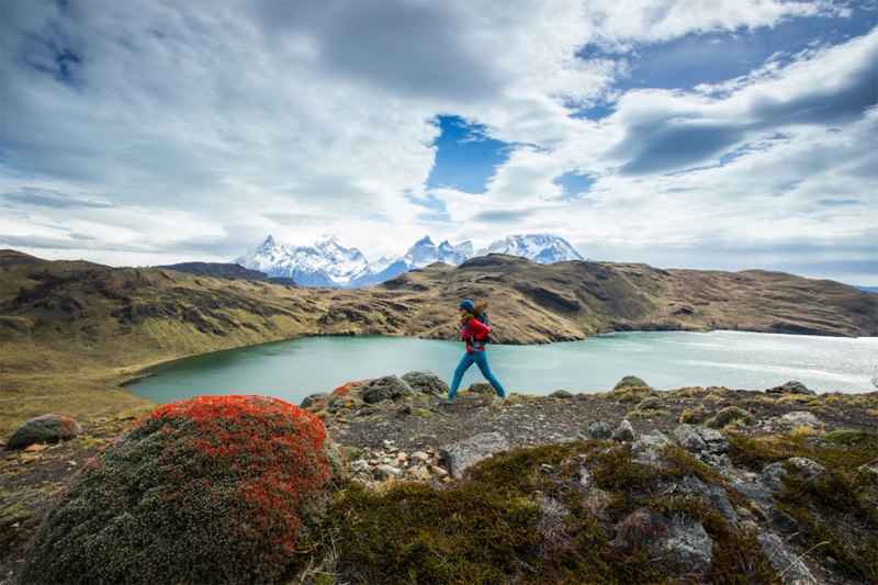 Hiker in a red coat walking with a backpack near a backcountry lake, mountains in the background.