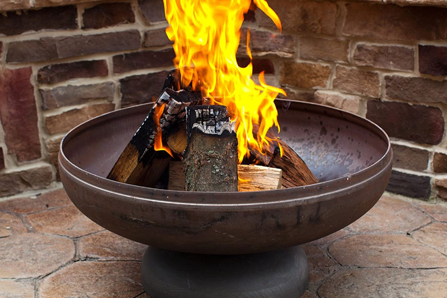 A fire burning in a round metal bowl on a stone patio against a brick wall.