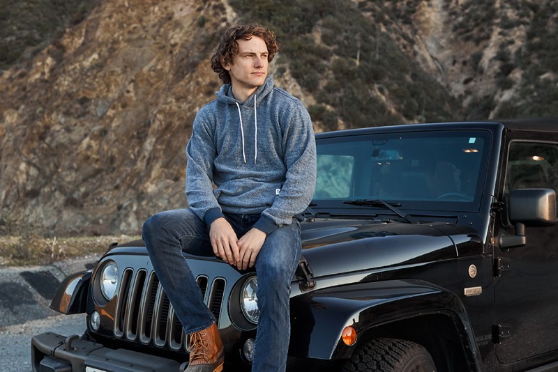 A man sitting on his car with a hoodie, staring off into the distance.