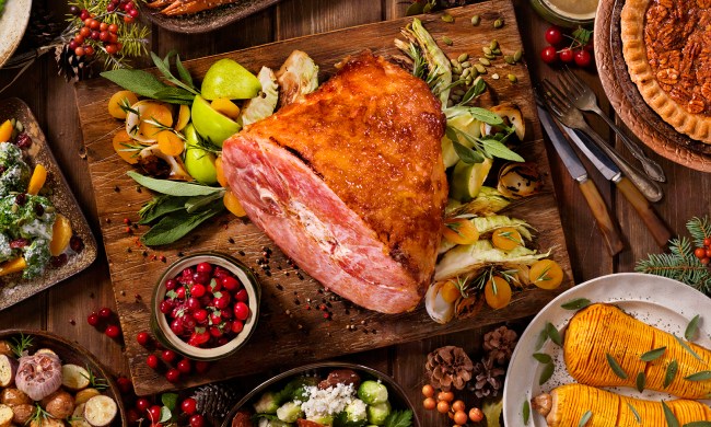 A large roasted ham on a wooden chopping board with assorted plates of food