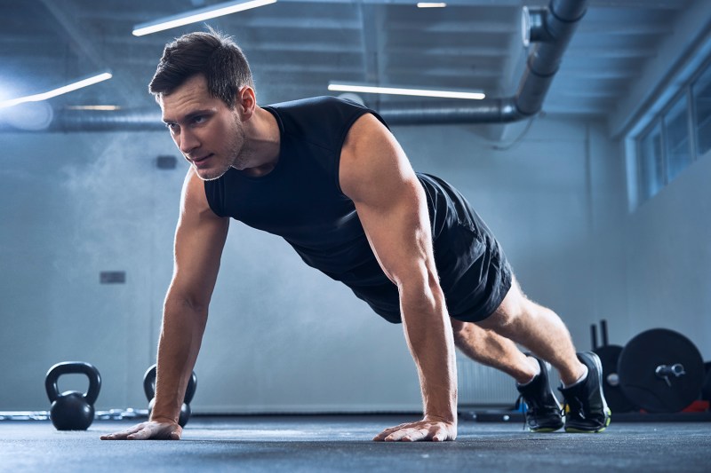 Lean yet muscular adult man wearing a black workout tank top as he completes pushups.