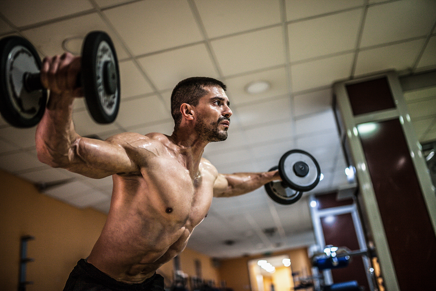 Shirtless man carrying two weights on each hand as he demonstrates the central back workout the Rear Flys.