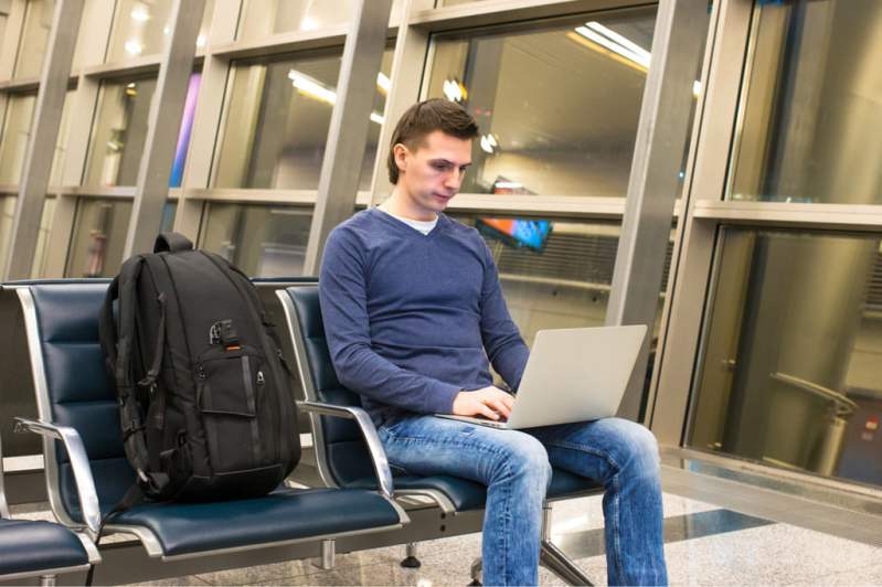 Man working on his laptop in the airport, seated next to a travel backpack.