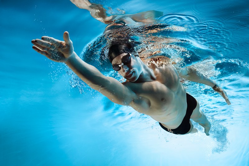 Man swimming while wearing a swim cap, goggles, and shorts.