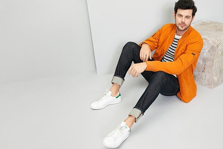A man seated against a stone block, modeling dark-wash jeans rolled at the cuffs, white sneakers, and an orange jacket over black and white striped shirt. 
