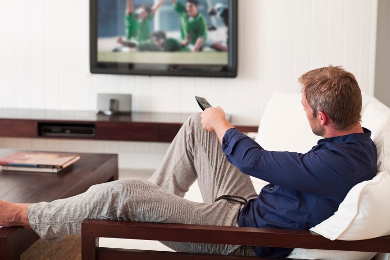 A man sitting on a sofa with his leg on the table is holding a remote control to change the channel.