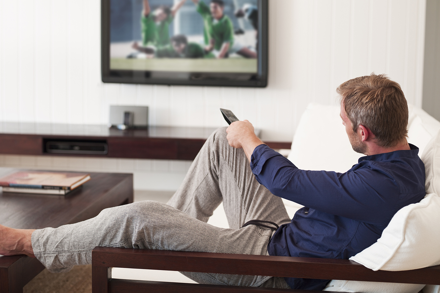 A man sitting on a sofa with his leg on the table is holding a remote control to change the channel.