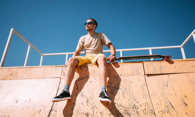 Young man with earbuds and longboard sitting on top of halfpipe in skatepark