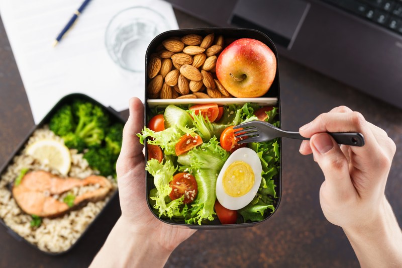 Photo of a man's hands holding a fork to eat from a bento box filled with salad, nuts, and an apple.