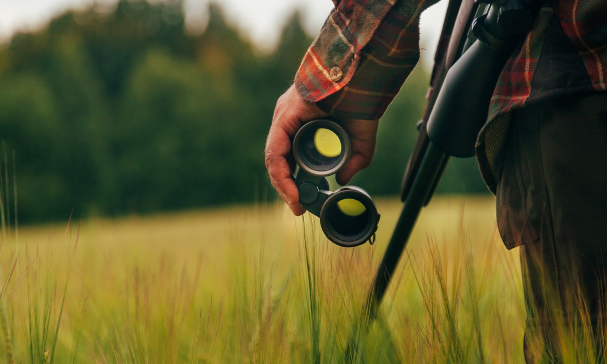 A person holding a pair of binoculars with his left hand in an open field.