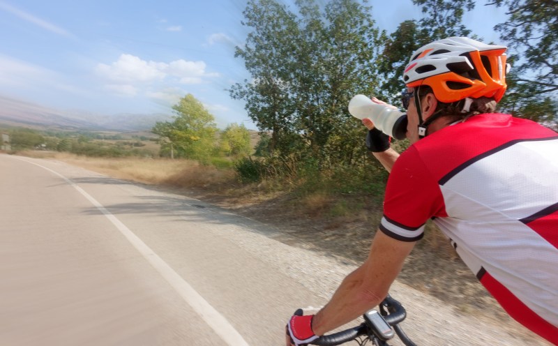 A biker drinking an electrolyte beverage from his water bottle while cycling on the street.