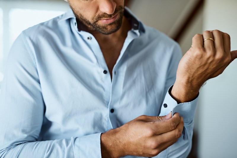 man with a beard buttoning a light blue dress down shirt with black buttons.