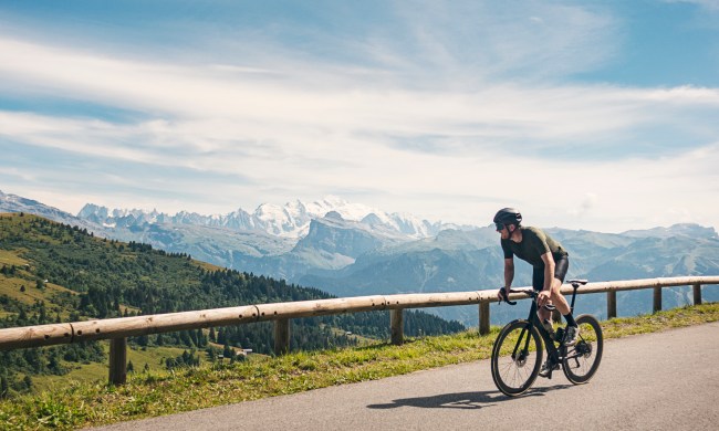 A man in helmet riding a bike on a road with railings with a view of a mountain and forest in the background.