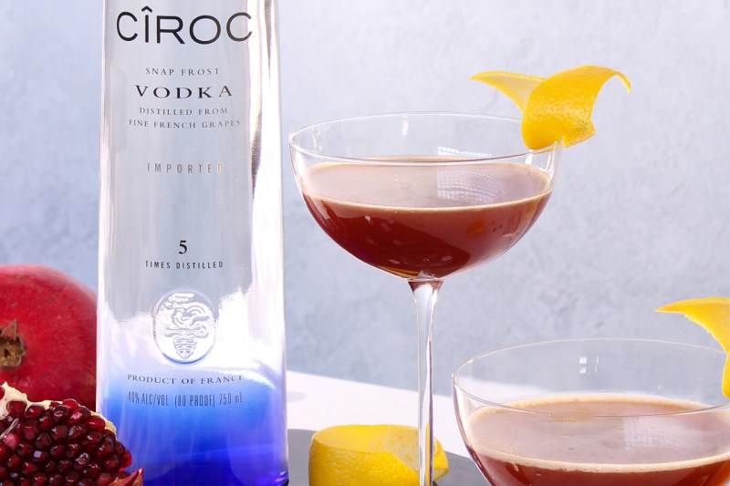 Two glasses of Ciroc Pomegranate Spring Fever Cocktail with lemon garnish beside a bottle of Ciroc Vodka and fruits.