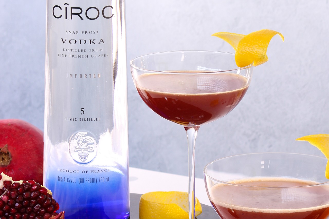Two glasses of Ciroc Pomegranate Spring Fever Cocktail with lemon garnish beside a bottle of Ciroc Vodka and fruits.