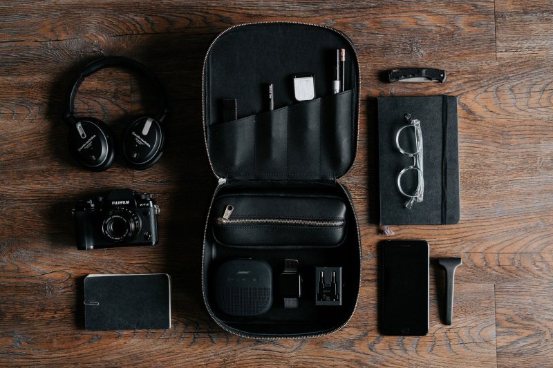 Flat-lay of all-black EDC (everyday carry) items, including headphones, a camera, glasses, a pocket knife, and more