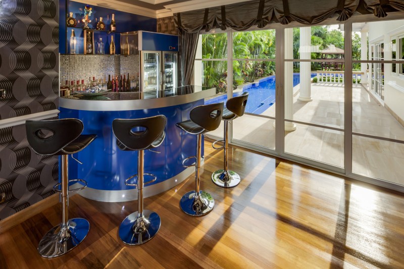 Home bar area with a blue-colored bar with bar stools next to floor to ceiling windows.
