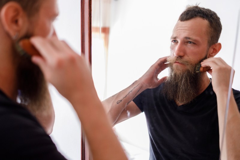 Man brushing beard with comb while looking at mirror in living room