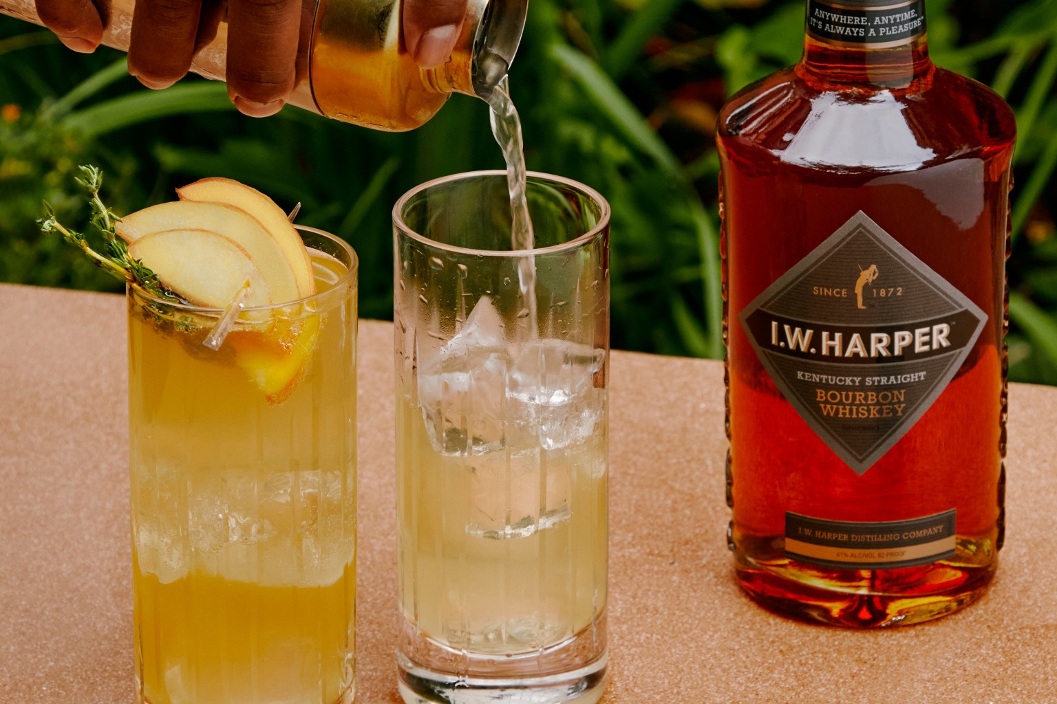 A Harper Spring Thyme being poured into a glass beside a glass of cocktail with fruit garnish and a bottle IW Harper Bourbon Whiskey.