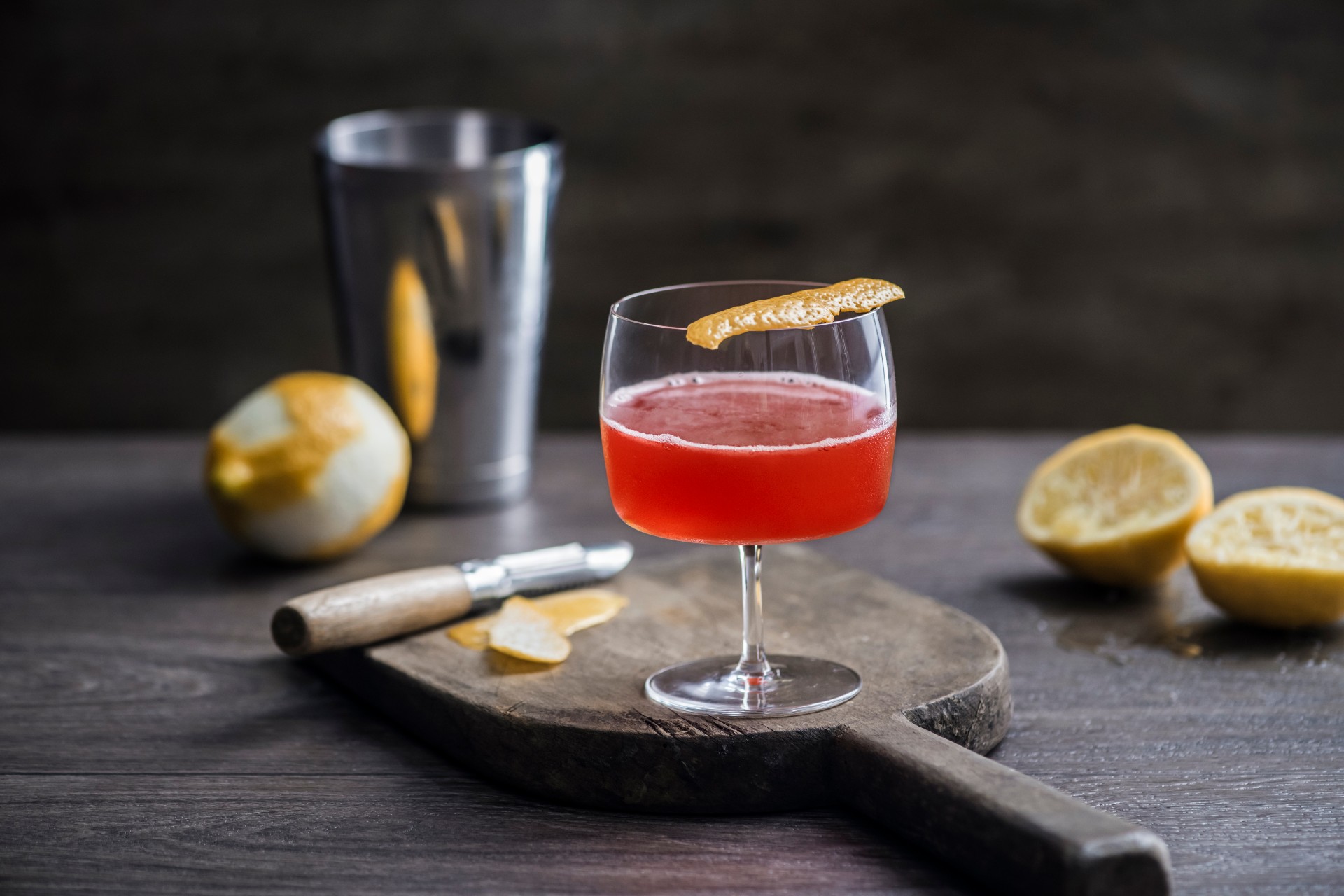 A Scofflaw in Love Cocktail with orange peel on top, served on a cutting board with a cocktail shaker and oranges in the background.