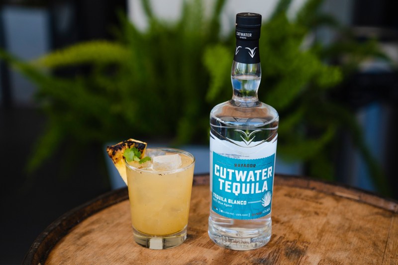 A bottle of Cutwater Tequila and a glass of Grill & Chill cocktail garnished with pineapple and leaves on a round wooden table