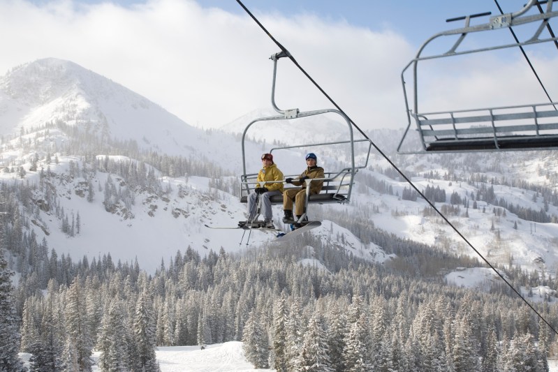 A skier and a snowboarder ride a ski lift with mountains and trees in the background.