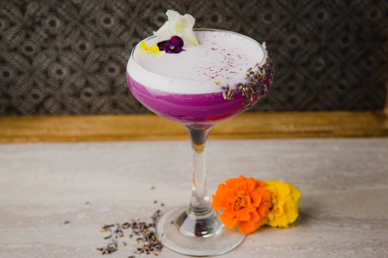 A Lavender Lasso cocktail with flowers and seeds beside it on a table