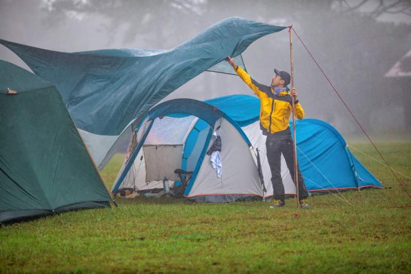 A man holds a pole and tries to control a tarp flapping over a tent in the wind and rain.