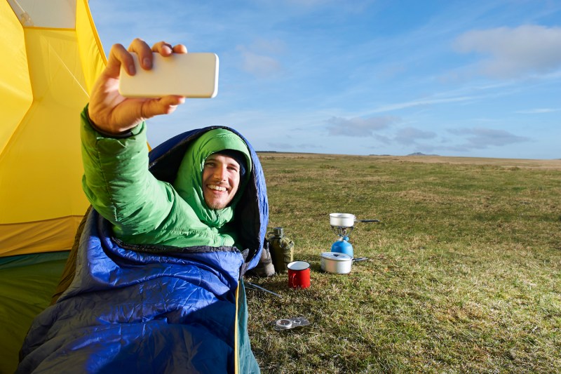 Camping man in a sleeping bag taking a selfie on a grass-covered hill.