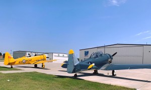 Two fighter planes, one yellow one black, sit facing an aircraft hangar.