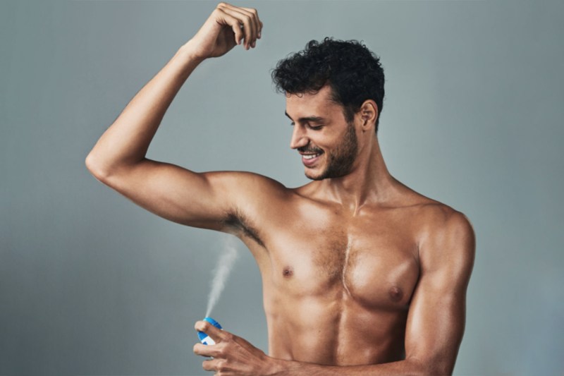 A shirtless man shown from the waist up, spraying a deodorizing body spray into his armpit.