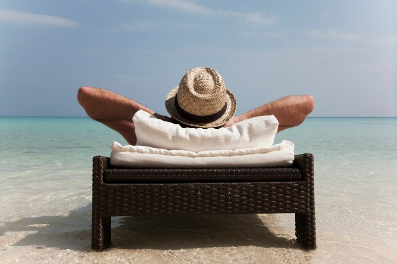 A man with his hands on the back of his head is wearing a straw hat and relaxing on the beach.