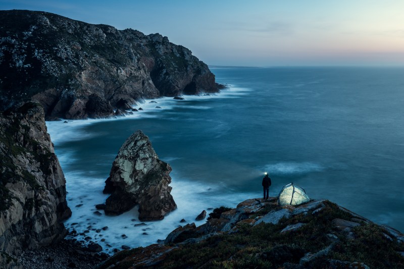 Camper standing on the edge of an oceanside cliff next to a lit-up geodesic tent.