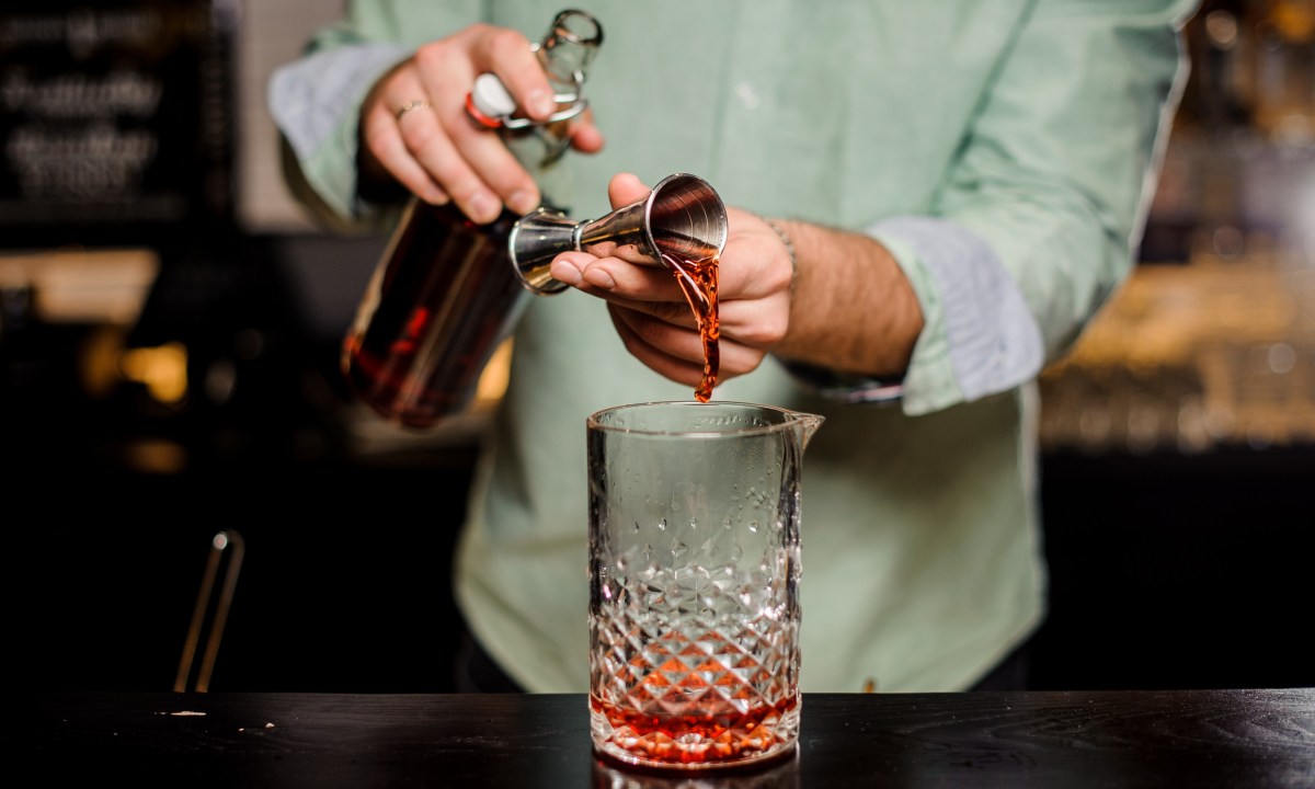 Bartender making alcoholic cocktail in red color, metal jigger and bar environment